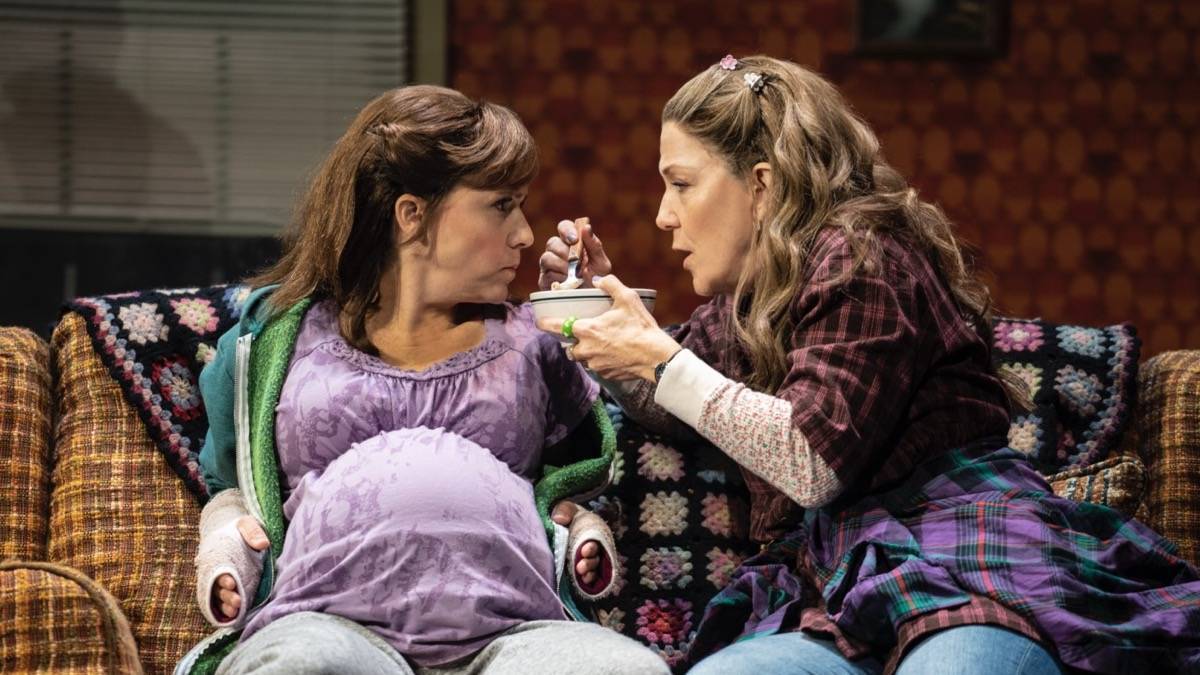 A woman holding a soup bowl attempting to feed another woman while sitting on a couch
